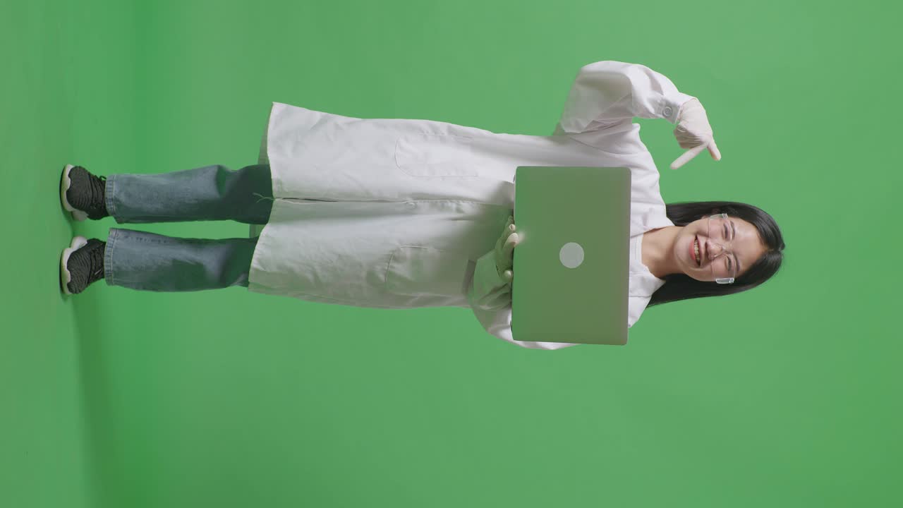 cuerpo lleno de mujer científica asiática sonriendo y señalando a una computadora portátil mientras estaba de pie en el fondo de la pantalla verde en el laboratorio