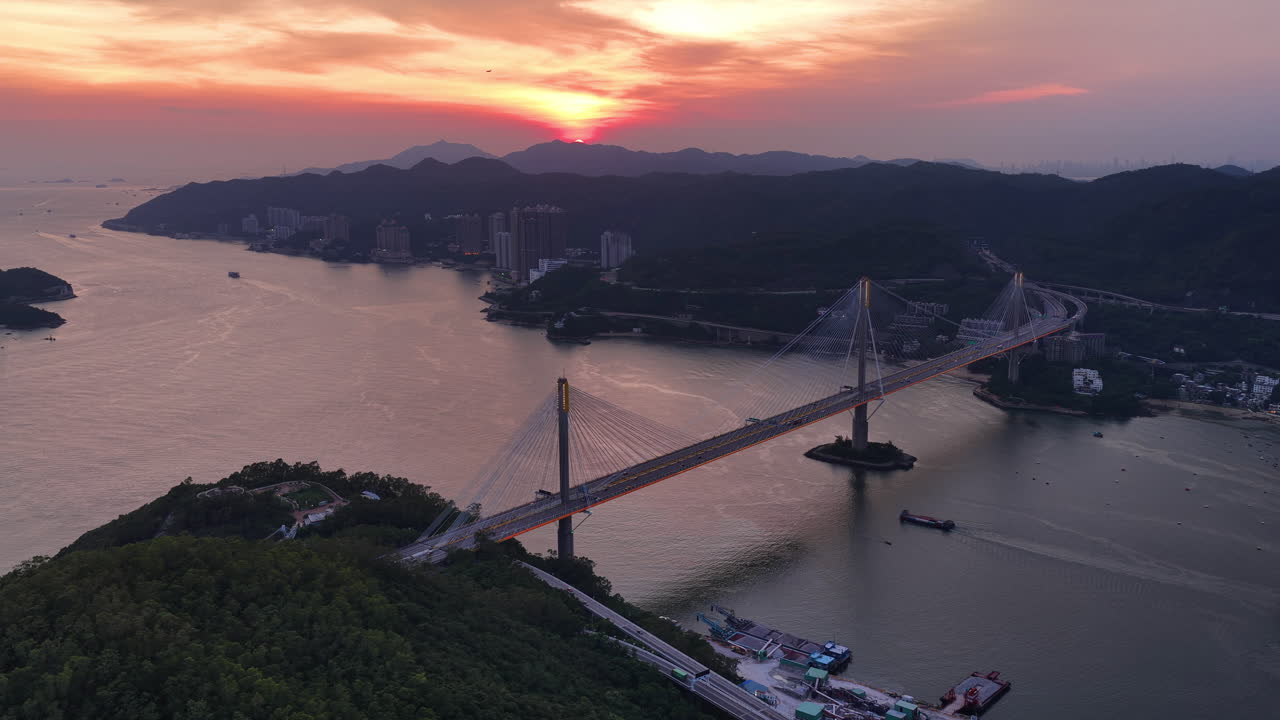 Cinematic Aerial view of Tsing Ma Bridge in Hong Kong at sunset, spanning over calm waters with city skyline, mountains, and ships in the distance, creating a serene urban and natural landscape scene