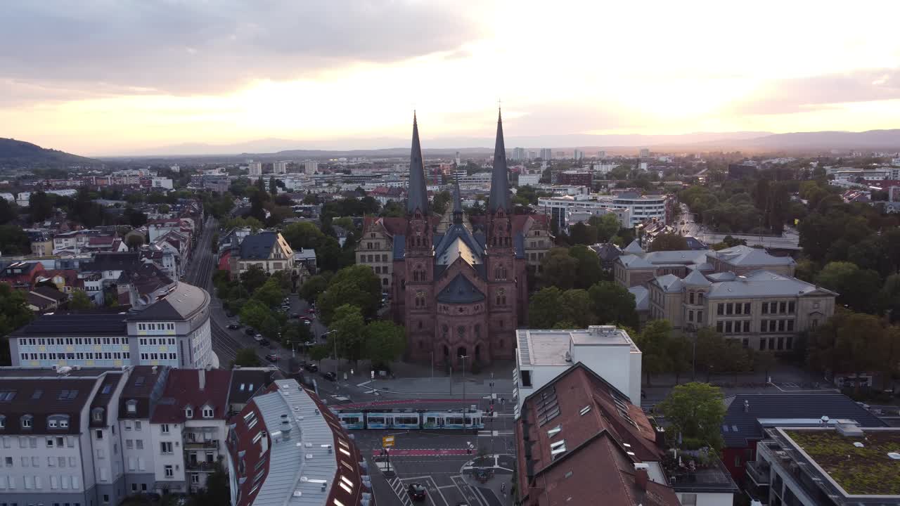 Drone flying away and revealing surroundings of catholic church Johanneskirche in gothic old town of Freiburg im Breisgau during sunset, Germany