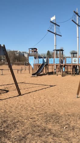 wide shot of an empty playground featuring a large pirate ship playset and swings on sandy ground under a clear blue sky.
