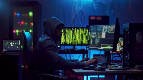 young male hacker in a hood working and hacking data at the two computers at one time while coding and tapping