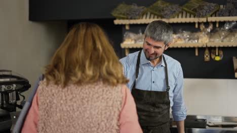 friendly smiling male cashier in apron giving paper bag with bread to the woman. female customer paying by phone and leave