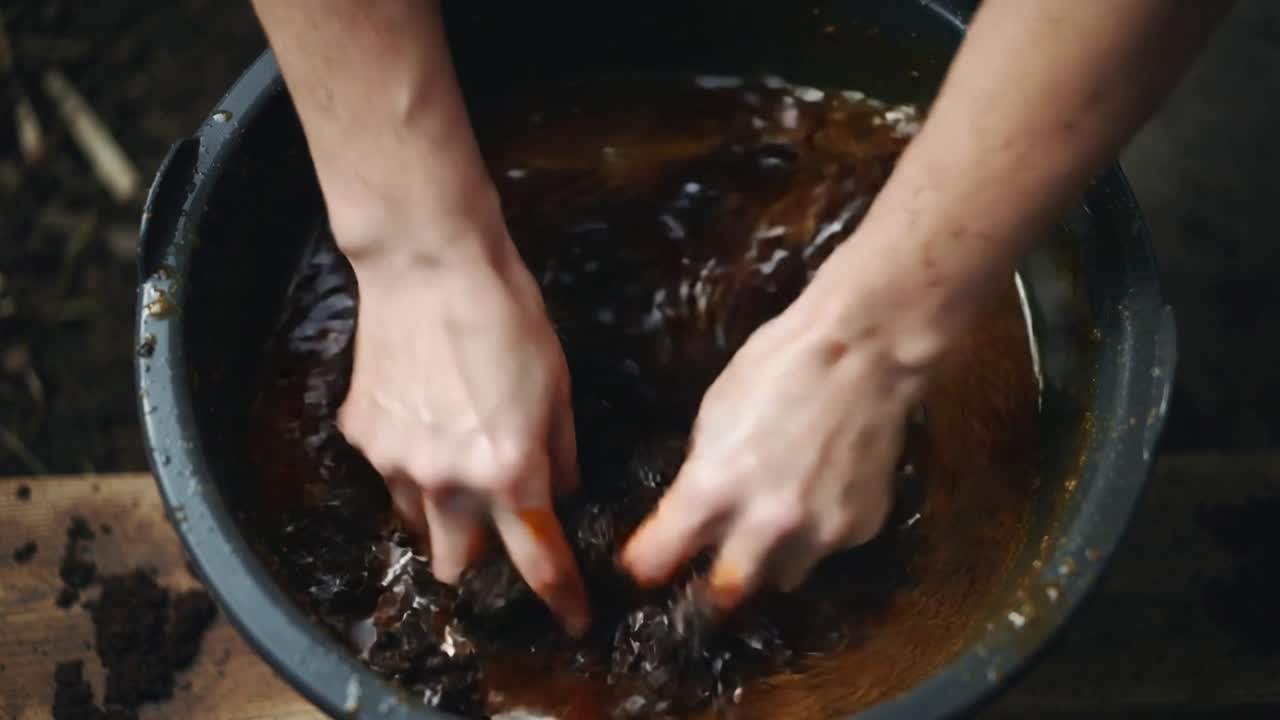 Mani nude che afferrano, impastano grumi scuri in un catino nero su una tavola, mescolando un liquido torbido.
