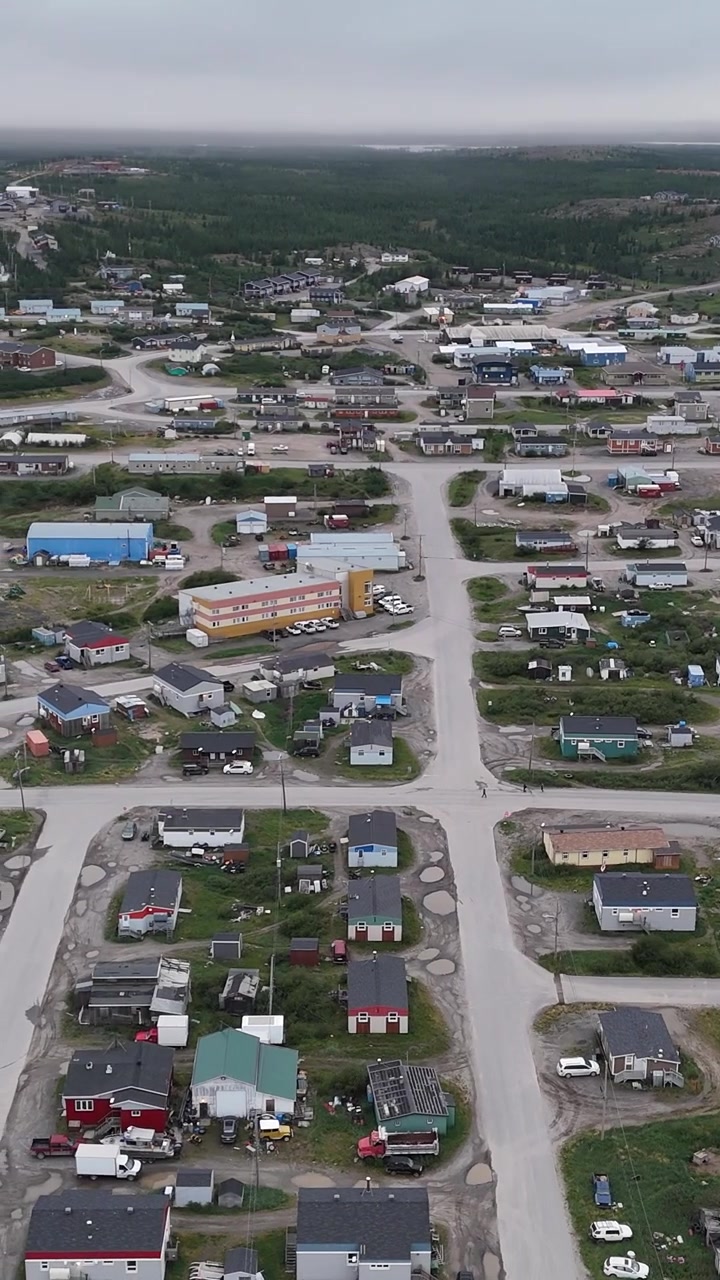 Vertical drone aerial slowly rotating left over houses, gravel roads, and tundra landscape in the town of Kuujjuaq, Nunavik, Quebec under an overcast summer sky.