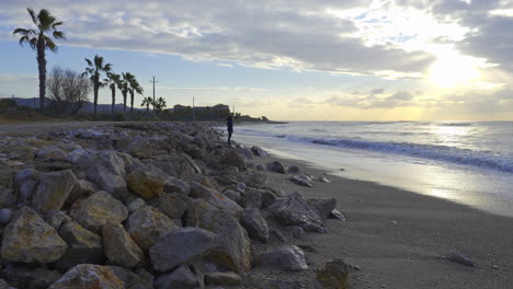 man-stands-on-rocky-ocean-front,-gazing-out-to-sea-as-morning-sun-breaks-through-clouds,-illuminating-picturesque-landscape