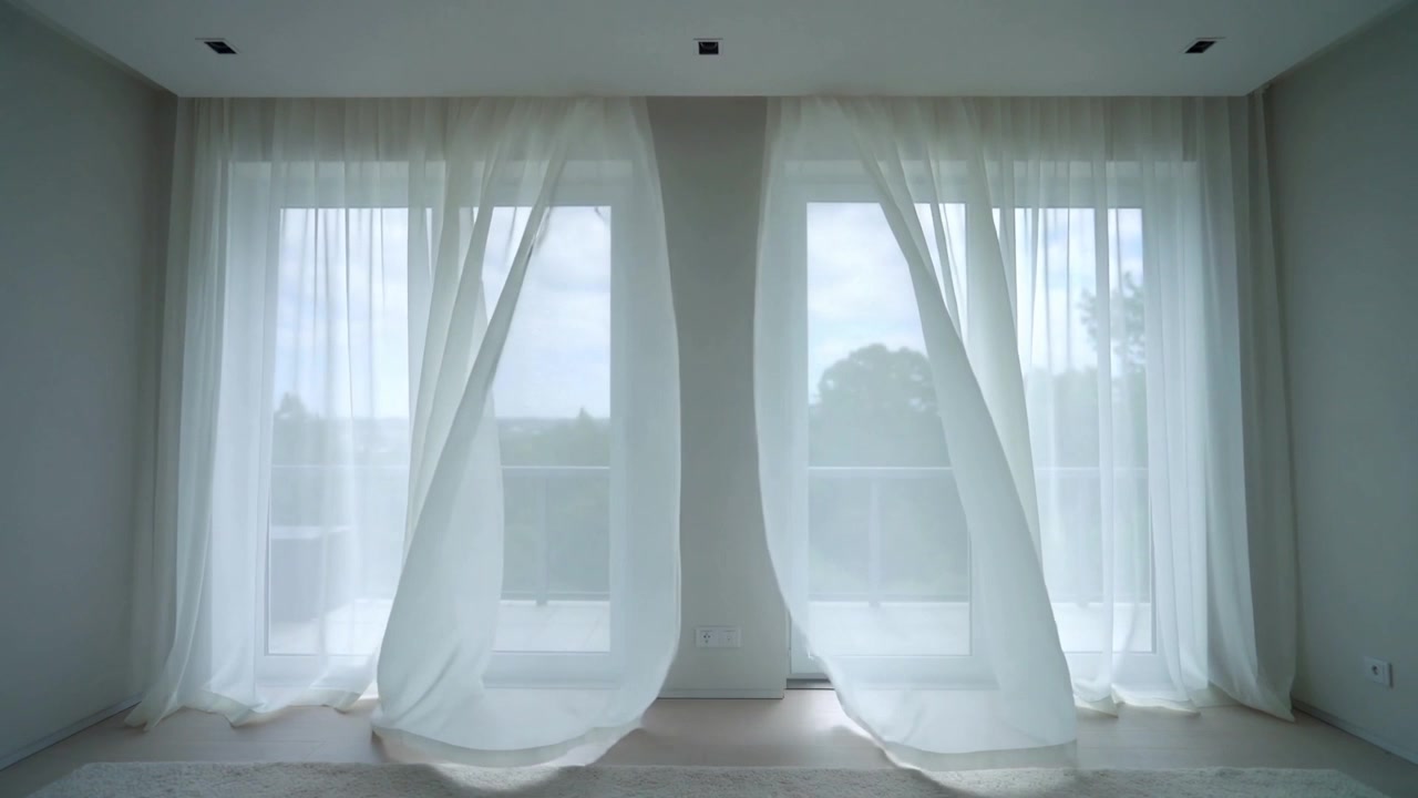 Tende velate che ondeggiano e si aprono verso l'interno per una leggera brezza in una stanza luminosa e vuota, rivelando un balcone