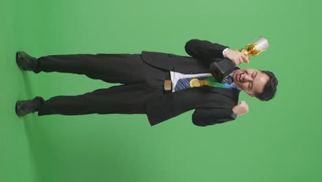 full body of asian business man in a suit and tie with a gold medal looking at a gold trophy then celebrating winning as the first winner on green screen background in the studio