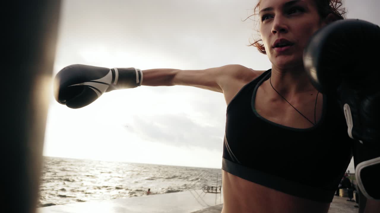 boxeadora dura con guantes golpeando una bolsa de pie contra el sol junto al mar. boxeadora entrenando temprano en la mañana en la playa. filmado en 4k