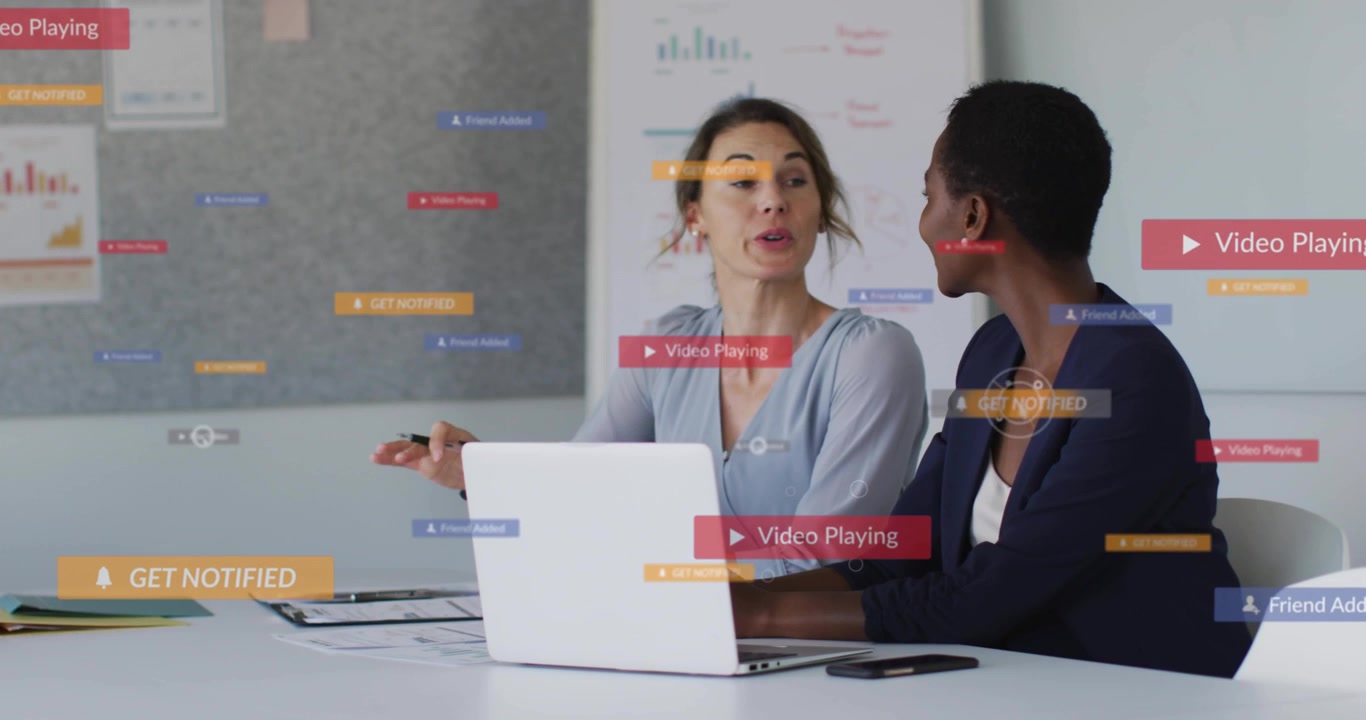 Two women presenting business papers in meeting, play badges appearing causing laptop reviewing