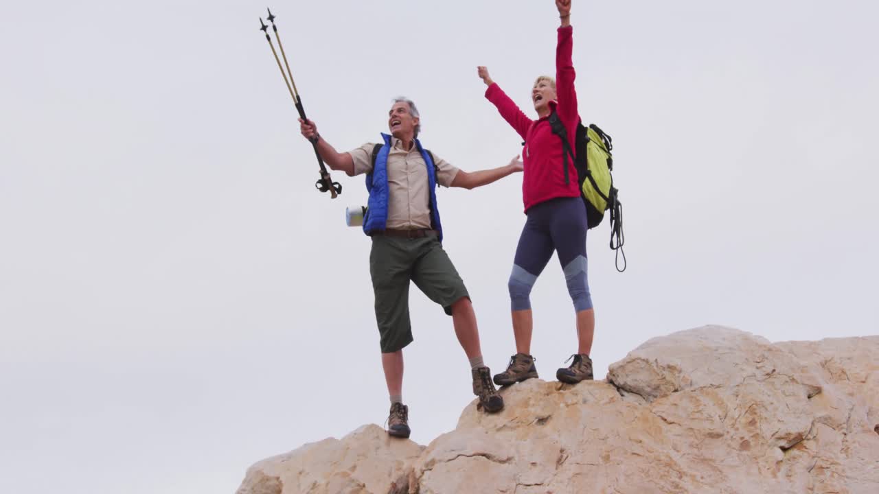 배 ⁇ 과 트레킹 기둥을 가진 고령 하이커 커플이 바위 위에 팔을 넓게 열고 서 있습니다.