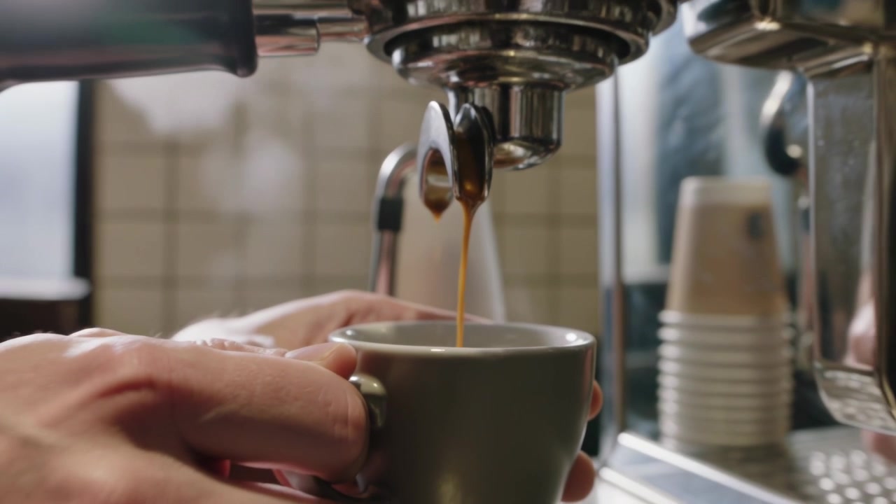 Macchina che eroga un espresso al bar, barista in camicia grigia che tiene ferma la tazza sotto i doppi beccucci, riempiendo la tazza.