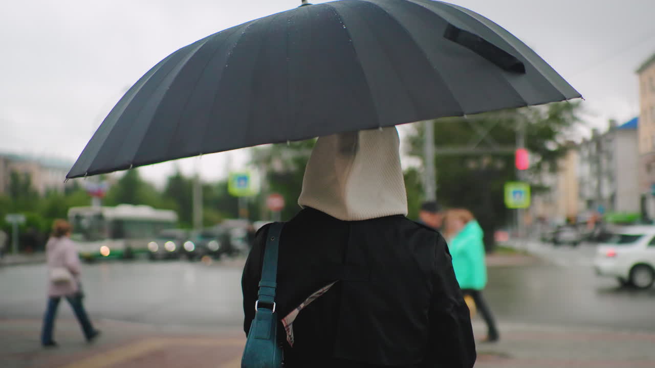 흐릿하게 보이는 보행자, 자동차, 건물이 배경에 있고, 우산을 쓰고 파란색 가방을 멘 후드를 입은 소녀가 비 오는 도시 거리를 걷고 있어요. 흐린 날씨에 도시의 라이프스타일을 보여주는 축축한 날씨의 통근 풍경이에요.