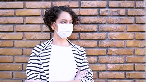 depressed young brunette woman with coronavirus mask leaning against the brick wall crosses her arms