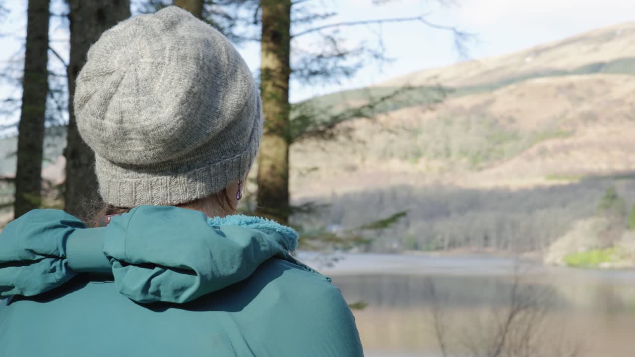 A camera looks over the shoulder of a woman eating her lunch while out for a walk in the forest, looking out across a Scottish Loch and hillside in the sun