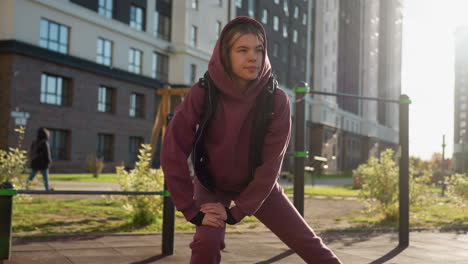 focused woman under sunny weather performing side lunge exercise with hands on legs on urban courtyard framed by autumn flowers and greenery, people in coats pass by against modern apartment backdrop