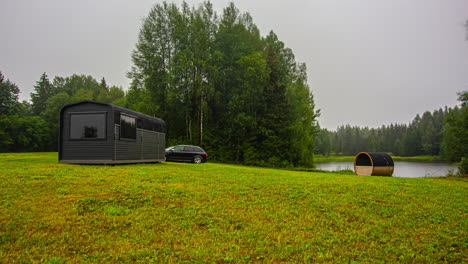 Time-lapse-shot-of-rainy-day-with-clouds-on-rural-grass-field-with-wooden-house-and-lake-in-background