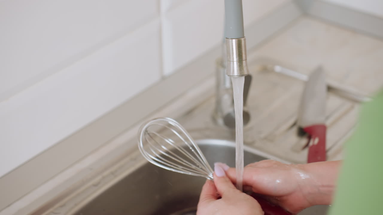 Close up of woman rinsing metal whisk under flowing kitchen tap water while holding knife beside sink, showcasing domestic cleaning task with clear view of hand