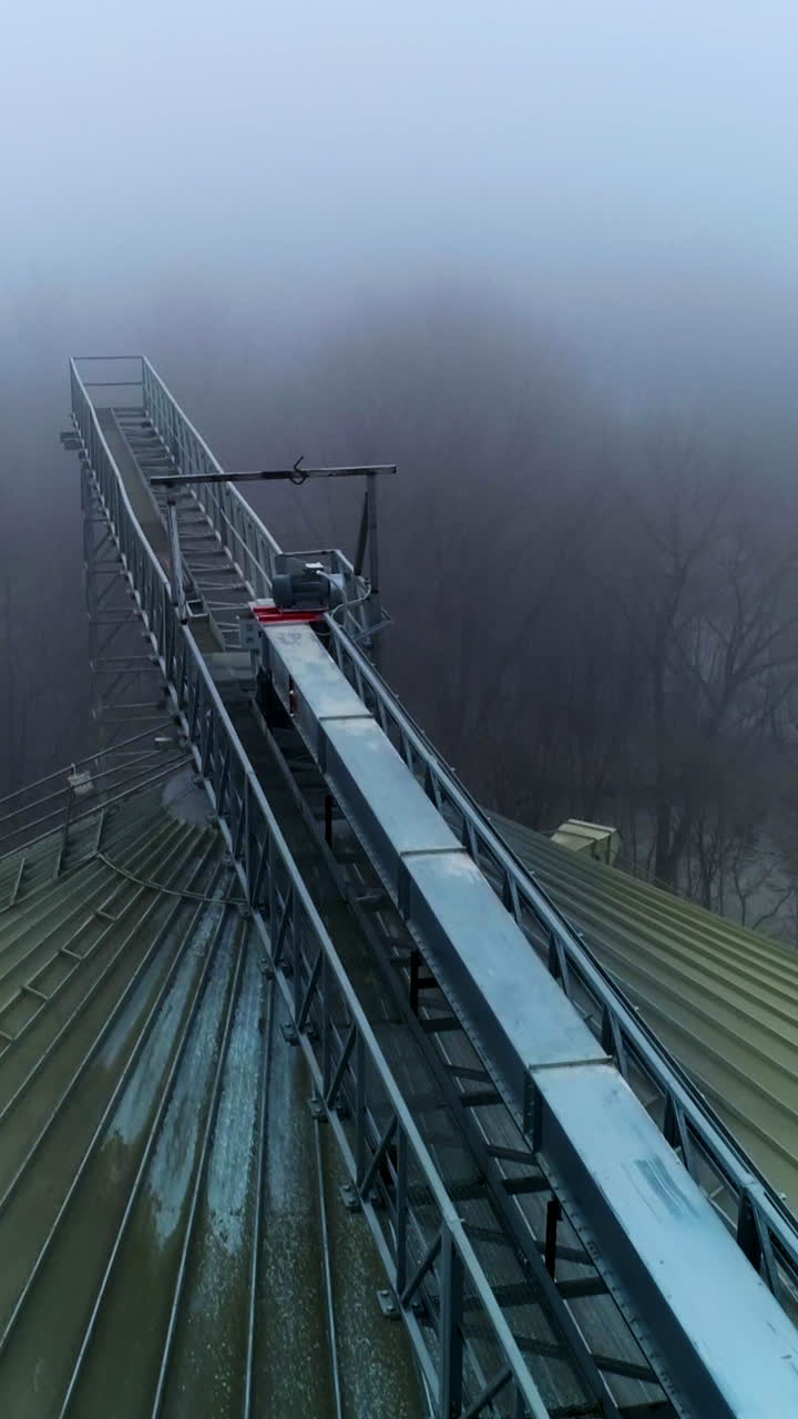 Big dirty rounded top of the granary tank. Flying along the metal support connecting the elevators. Top view on the winter foggy forest. Vertical video