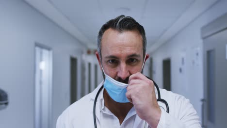 Portrait-of-smiling-caucasian-male-doctor-putting-on-face-mask-in-hospital-corridor