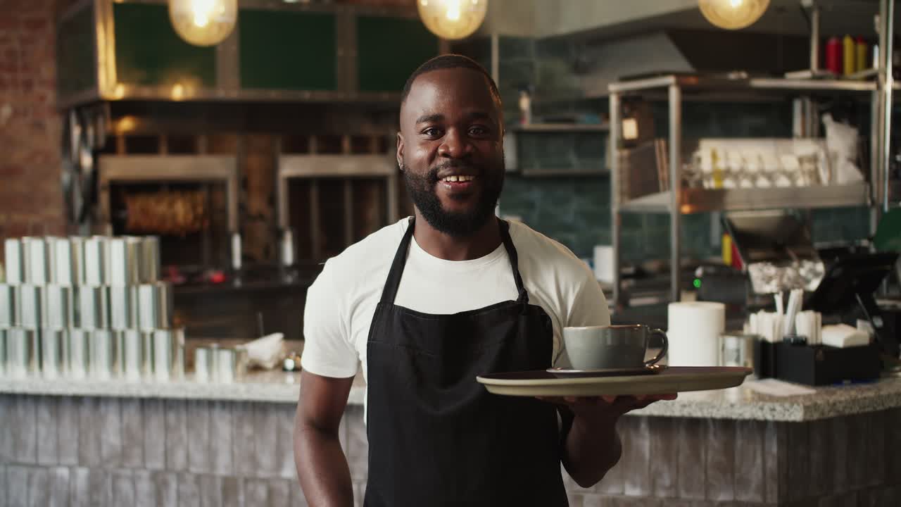 A Black person in an apron holds a tray with a cup of coffee and poses against the background of the doner market