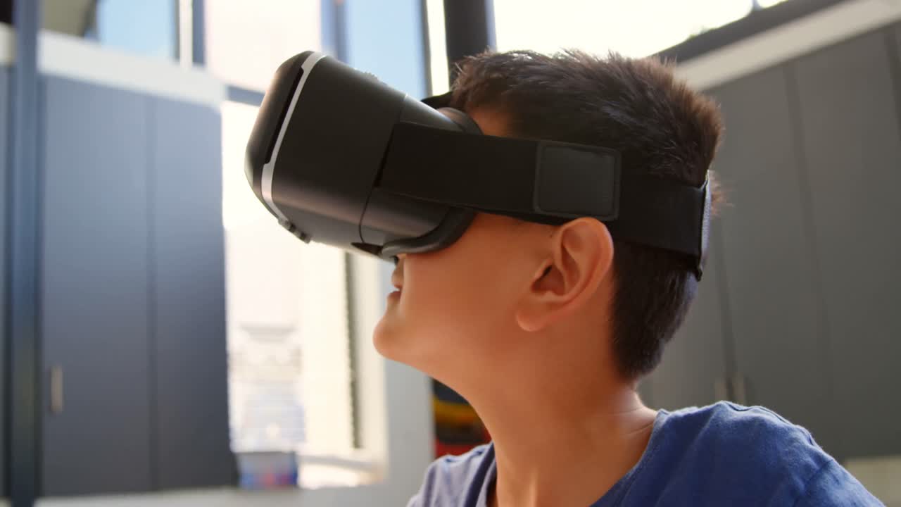 vista frontal de un escolar asiático sentado en un escritorio y usando auriculares de realidad virtual en el aula 4k