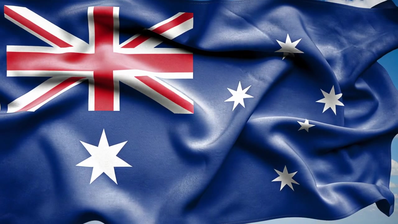 Bandera australiana ondeando revelando la Union Jack y las estrellas en el mástil contra el cielo azul, el viento impulsando su despliegue.