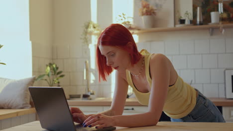 una joven pelirroja enviando mensajes de texto en una computadora portátil en una cocina moderna y soleada.