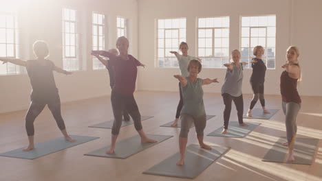 yoga-class-of-healthy-mature-women-practicing-warrior-pose-enjoying-morning-physical-fitness-exercise-workout-in-studio-at-sunrise