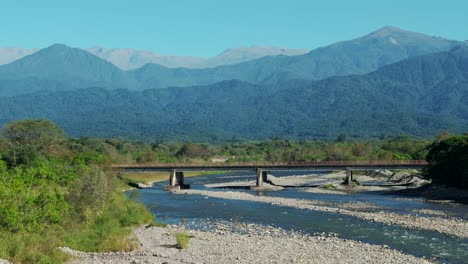 Side-panning-view-of-a-bridge-with-a-clear-river-and-the-stunning-Andes-Mountains-in-the-background,-in-northern-Argentina