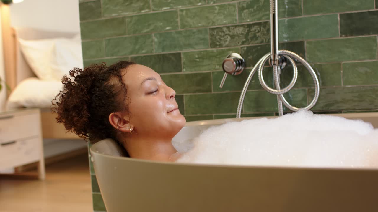 Relaxing in foam, African American woman scooping bubbles in freestanding tub by green tile wall
