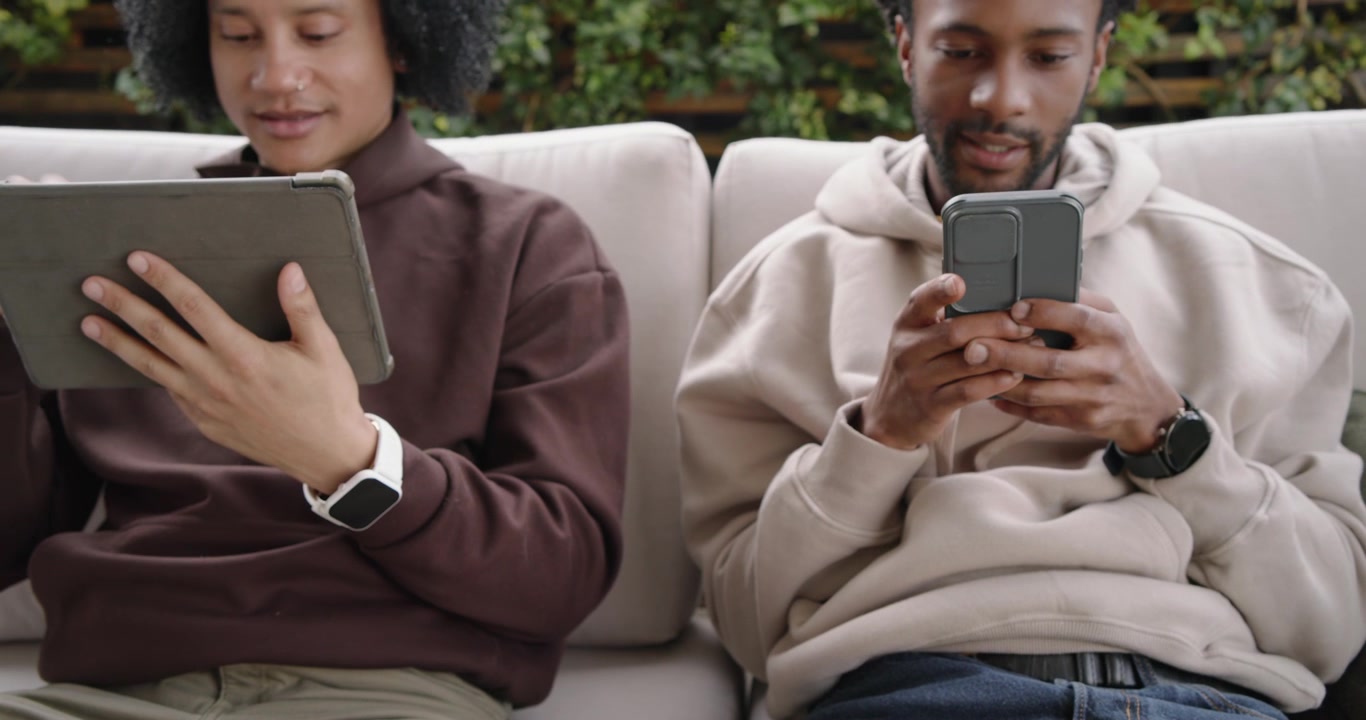 Diverse friends on patio sofa, woman browsing blue phone, man adjusting tablet, camera pulling back