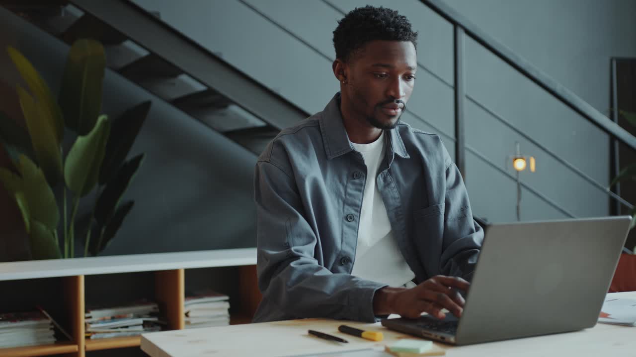 Young Black Man Typing on Laptop at Desk in Modern Office