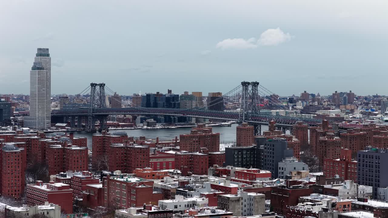 Veduta aerea invernale di New York City con il Ponte di Williamsburg che attraversa l'East River sopra edifici residenziali coperti di neve e densi isolati urbani. Ampia inquadratura panoramica. Giornata invernale nuvolosa