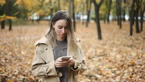 Young-blonde-woman-writing-message-on-cell-phone-in-a-autumn-park