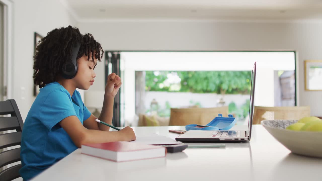 Happy biracial boy sitting at table using laptop and having online classes