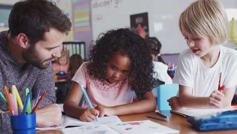 male elementary school teacher sitting at desk helping pupils in classroom