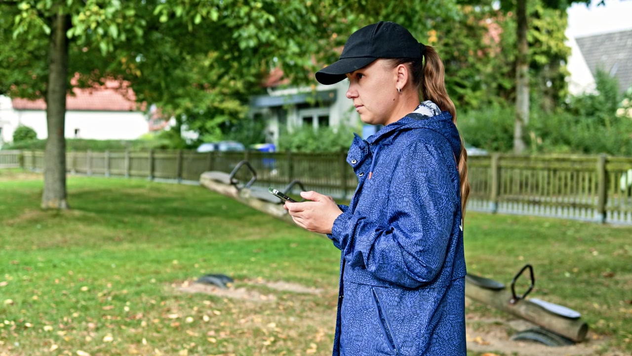 Woman stands in a park looking at her phone near playground equipment on a sunny day while wearing a blue jacket and a cap