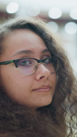 mujer joven con cabello rizado y gafas de pie en un centro comercial bien iluminado, mirando hacia la cámara con una sonrisa suave y suave, el fondo cuenta con una hermosa iluminación bokeh