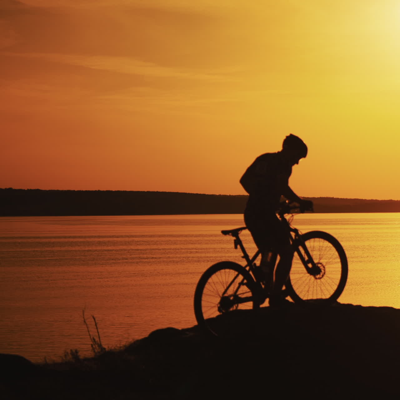Amazing view of a golden sunset path over water surface and a biker. Silhouette of a male biker in helmet rides up to the river during the setting sun.