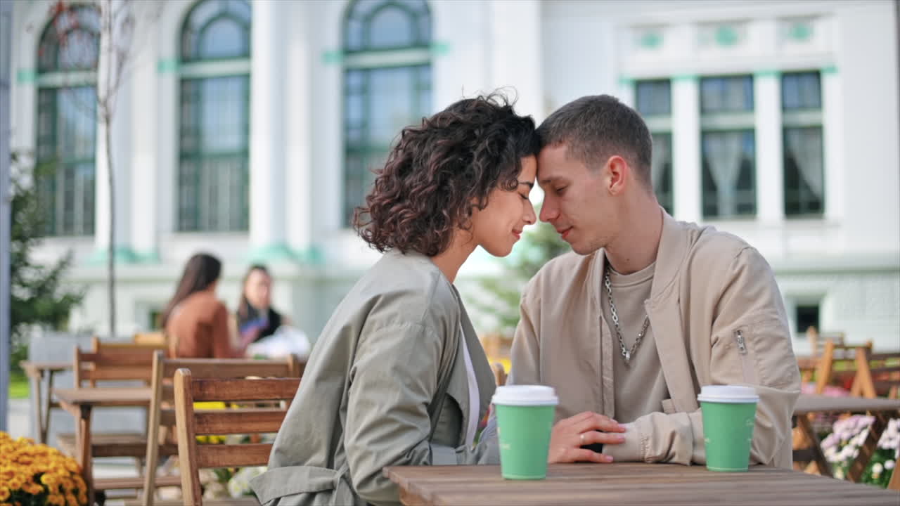 Una coppia romantica all'aperto vicino a un bar. Si tengono per mano, sorridono a occhi chiusi, caffè sul tavolo. Atmosfera autunnale. Slow motion