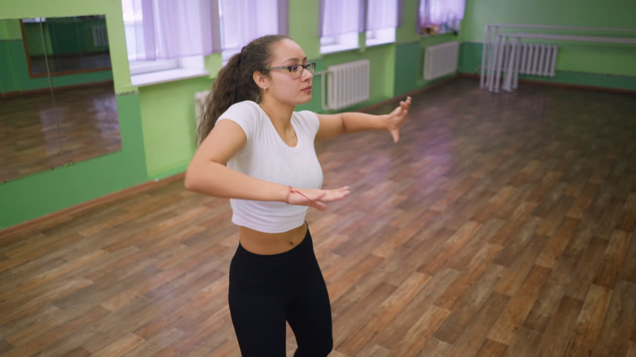 Young lady in white top black leggings and eyeglass dancing in room, mirror behind showing reflection, wooden floor and green painted walls enhancing energetic pose with graceful body movement