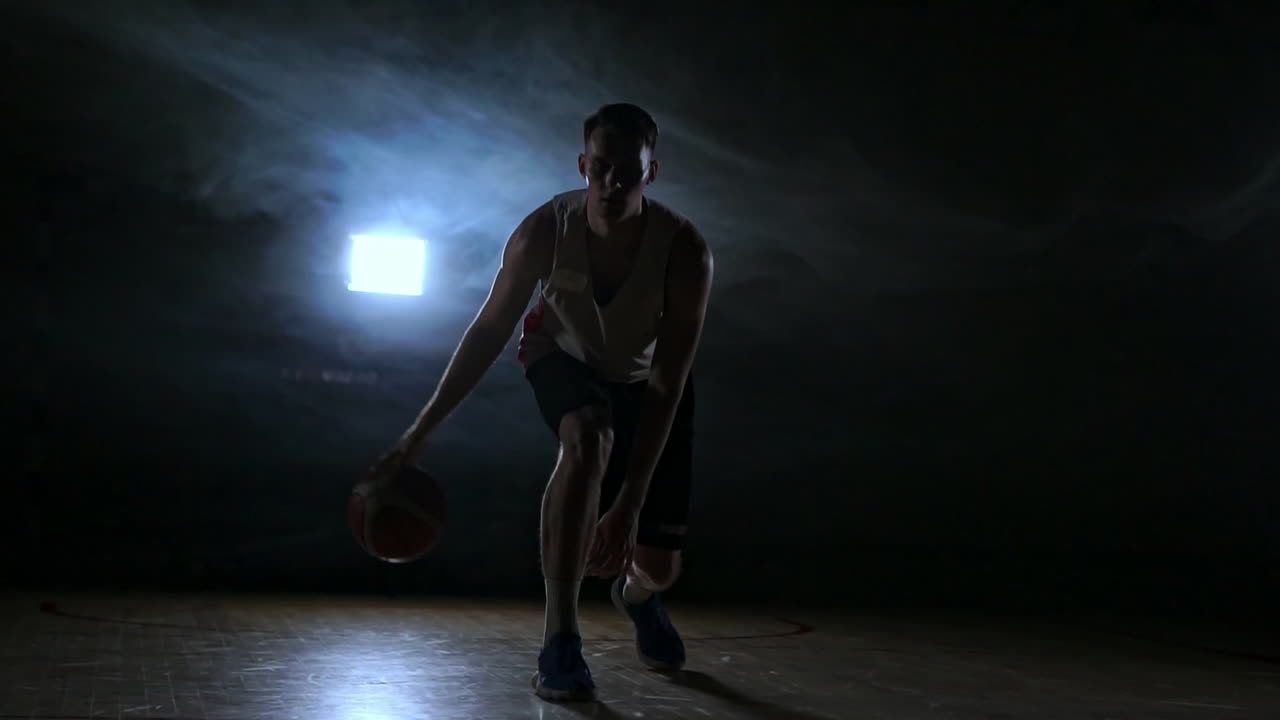 un joven adulto, jugador de baloncesto, pelota de dribble, cancha de baloncesto de interior oscura. cámara lenta