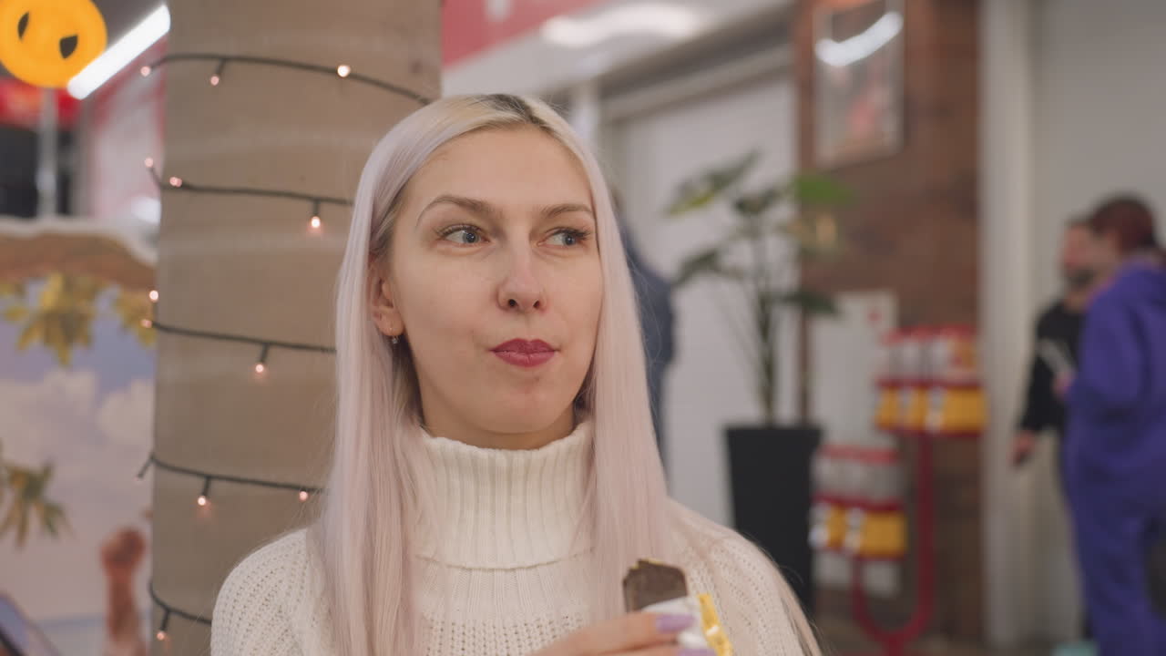 portrait of mindful woman biting chocolate bar in busy mall corridor, closeup of hand holding wrapper and lips tasting sweet treat, blurred shoppers passing by, retail interior