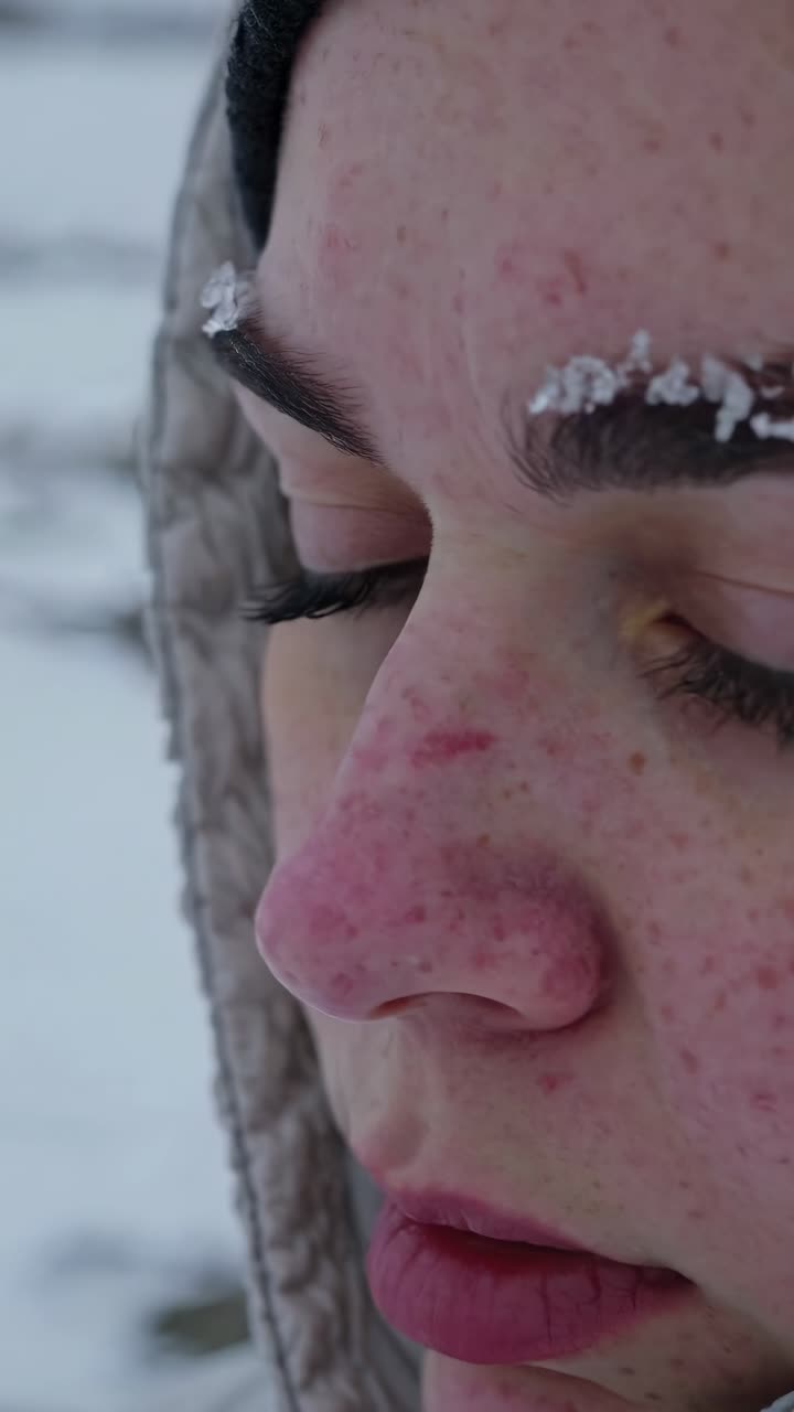 Snowflakes delicately rest on the eyebrow of a young woman braving the cold, her face touched by the winter chill as she experiences the serene beauty of a snowy landscape