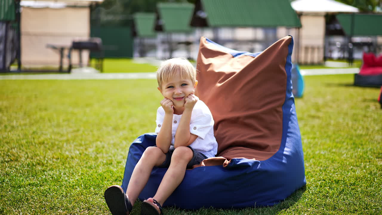 Smiling beautiful kid sits in a comfortable bean bag chair outdoors. Approaching blond boy spending time in resort zone.