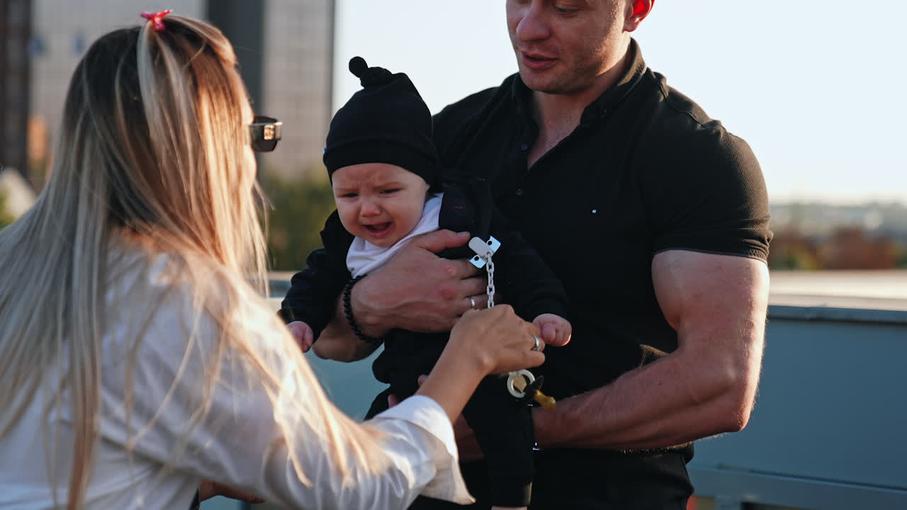 Muscular Caucasian man wearing black t-shirt holding a crying baby. Long-haired blonde woman tries to calm the infant giving him a pacifier. Family outdoors.