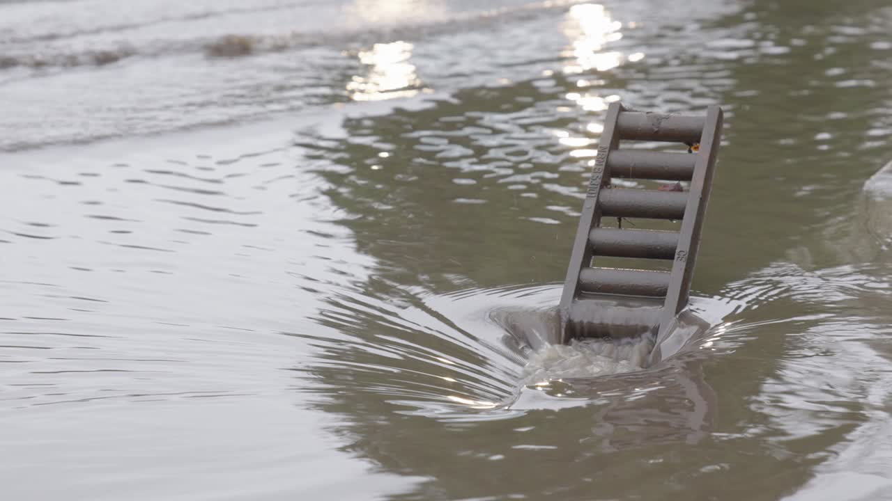 Reflections Of Cars Passing By A Flooded Street With Rainwater Flowing Into Drainage Hole In The Gutter
