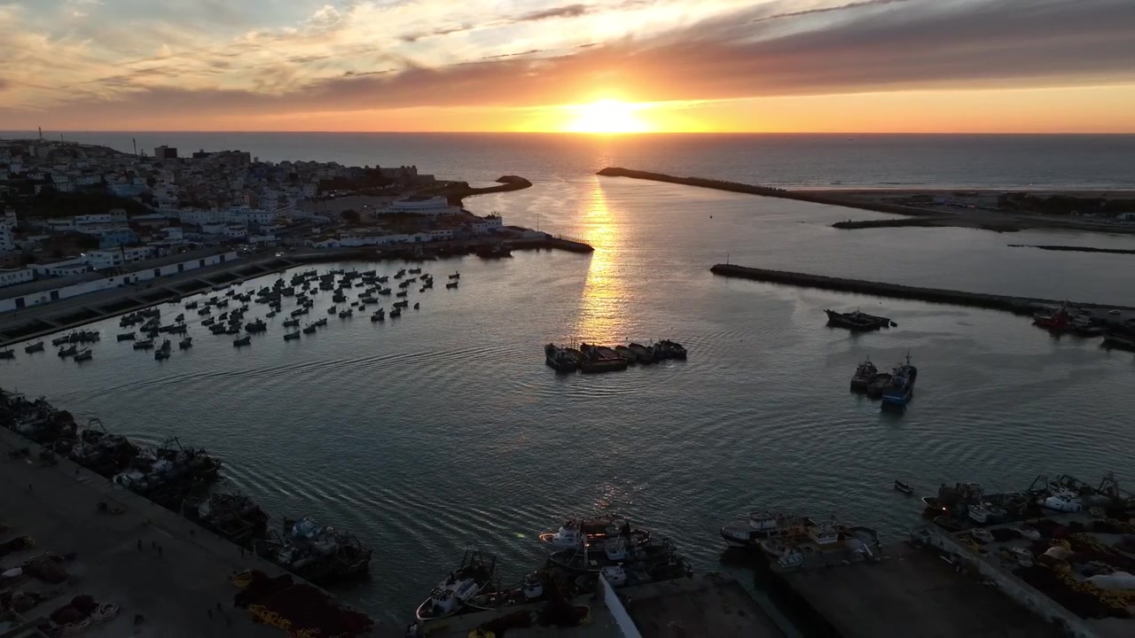Vista aérea de uma frota de pesca dentro do porto de Asilah ao pôr do sol, com pequenos barcos espalhados pela água calma, embarcações maiores perto do cais e luz dourada do Atlântico refletindo por todo o porto