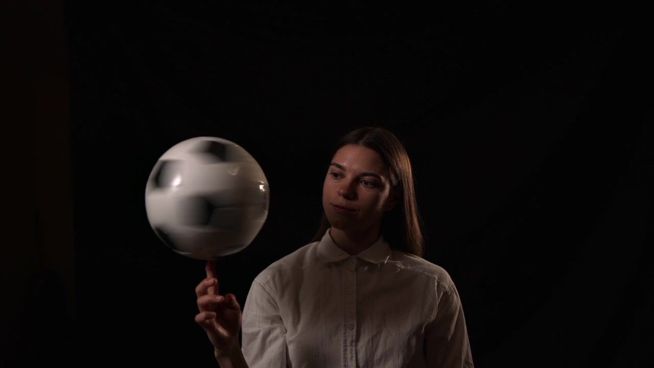 Giovane donna in camicia bianca si protende verso l'alto verso una sfera metallica lucida su uno sfondo scuro, con forte contrasto e un'atmosfera da studio minimalista e suggestiva.