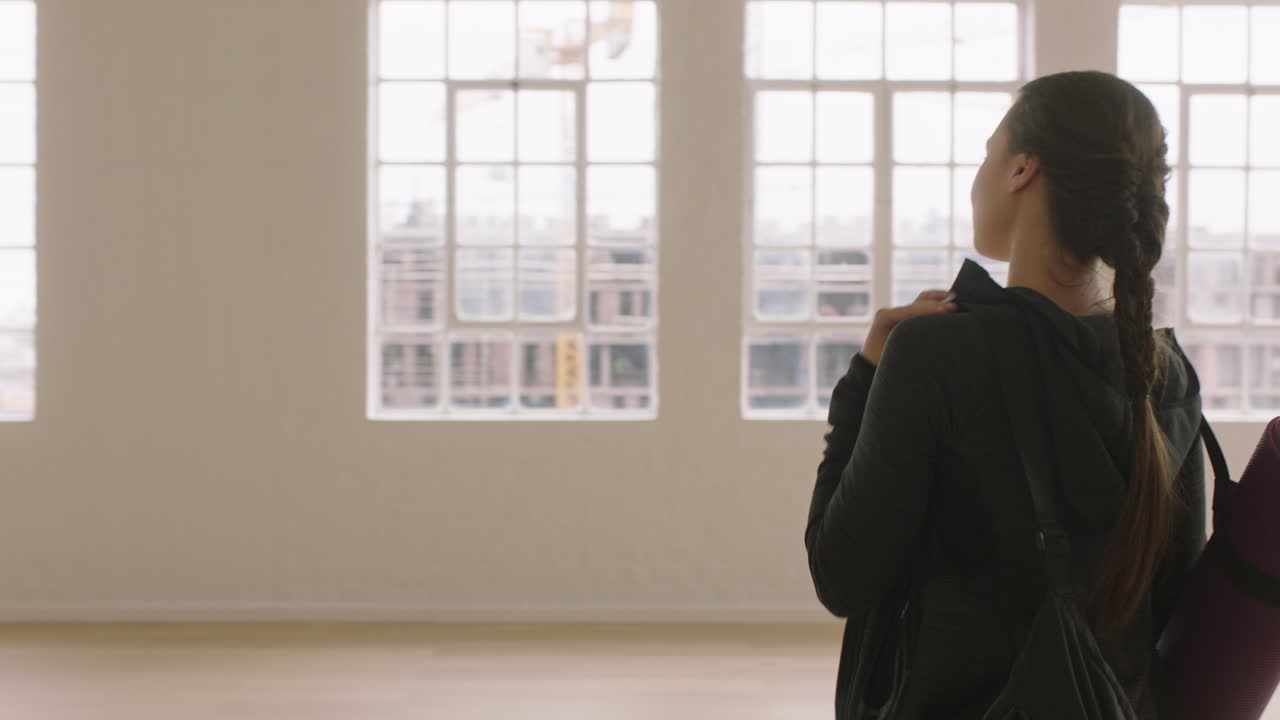 아름다운 요가 여성이 운동 스튜디오에 들어가 운동 준비를 하고 창밖을 내다보며 건강한 라이프스타일을 즐기며 미리 계획을 세웁니다.